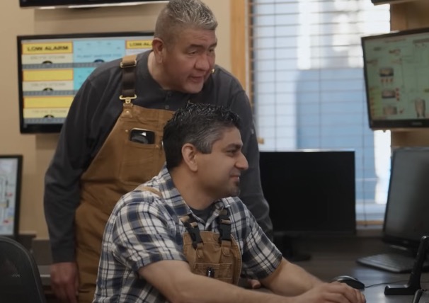 A factory worker sits while looking at a screen, with a colleague behind him, standing.